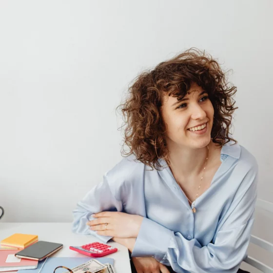 Smiling woman with curly hair sits at a desk in a light blue blouse, with cash, a calculator, glasses, and a smartphone on the desk in front of her.