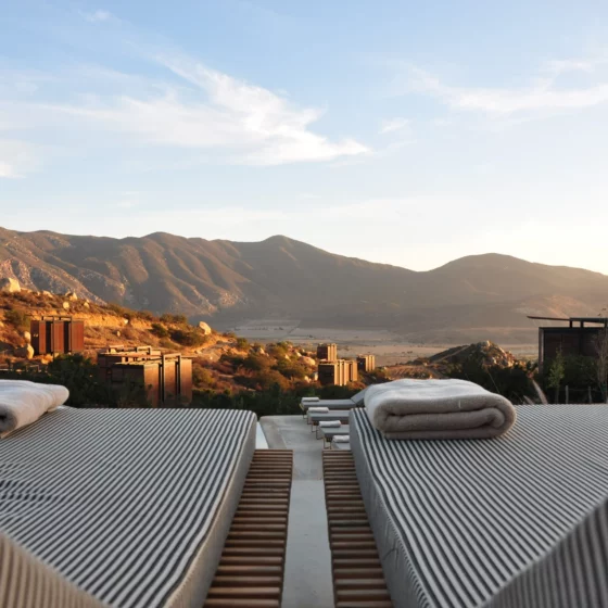 Row of striped lounge chairs with folded towels by a pool, mountains in the distance at sunset.