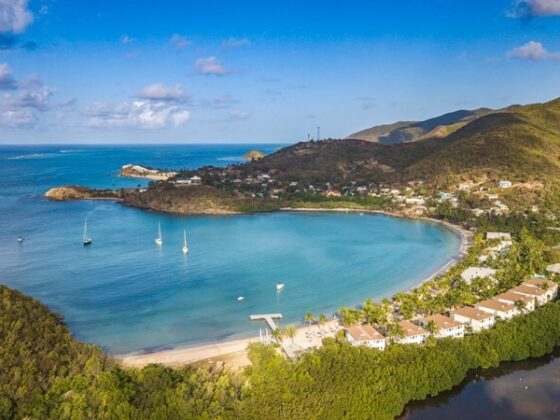 Aerial view of a crescent bay with turquoise water, several boats, a sandy beach, and rows of white-roofed houses along the shore. Green hills rise in the background.