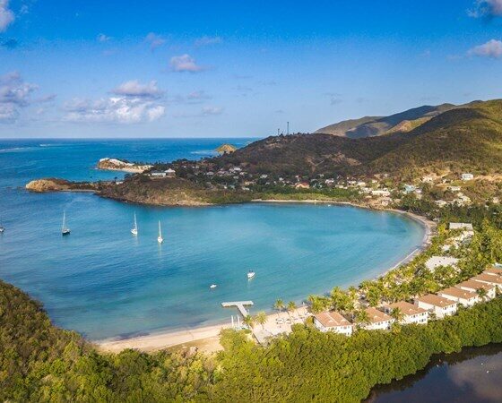 Aerial view of a crescent bay with turquoise water, several boats, a sandy beach, and rows of white-roofed houses along the shore. Green hills rise in the background.