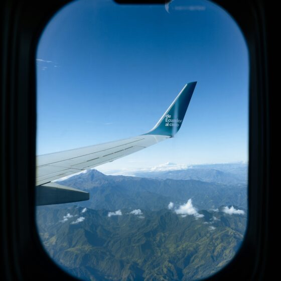 Wing of an airplane against a clear blue sky, with a mountain landscape below, seen from a window in flight.