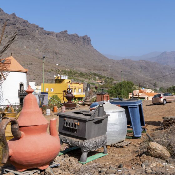 Old windmill in a desert yard surrounded by large ceramic pots and scattered metal containers, mountains in the background.