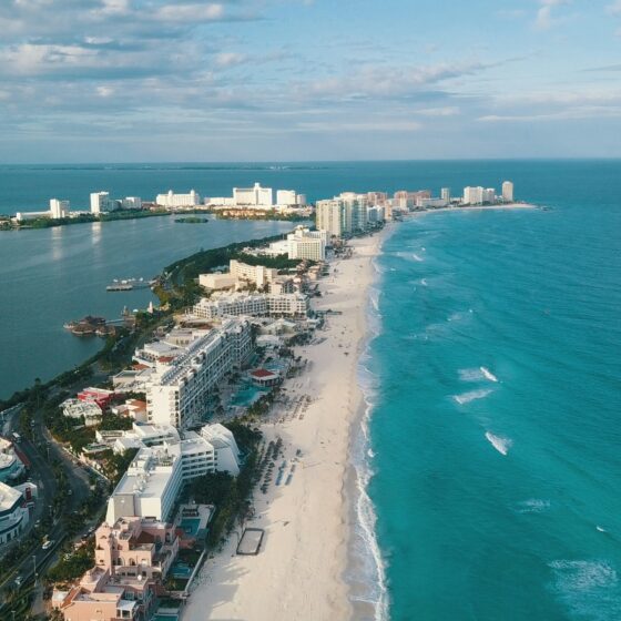Aerial view of a coastal resort with white hotels lining a long white-sand beach along turquoise water and a narrow barrier island.