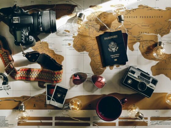 Flat lay of travel gear on a world map: camera with strap, passport, string lights, sunglasses, polaroid photos, a mug, and a vintage radio.