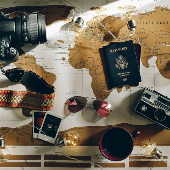Flat lay of travel gear on a world map: camera with strap, passport, string lights, sunglasses, polaroid photos, a mug, and a vintage radio.