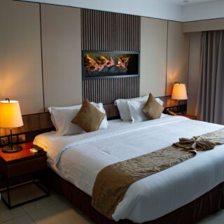 Hotel room featuring two large beds with white linens, beige pillows, and a shared dark wood headboard with vertical slats, flanked by bedside lamps.