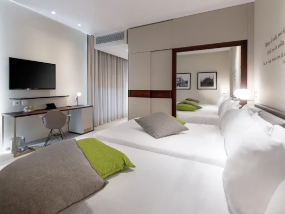 Hotel room with two twin beds in the foreground, gray pillows and green cushions, and a desk with a wall-mounted flat-screen TV.
