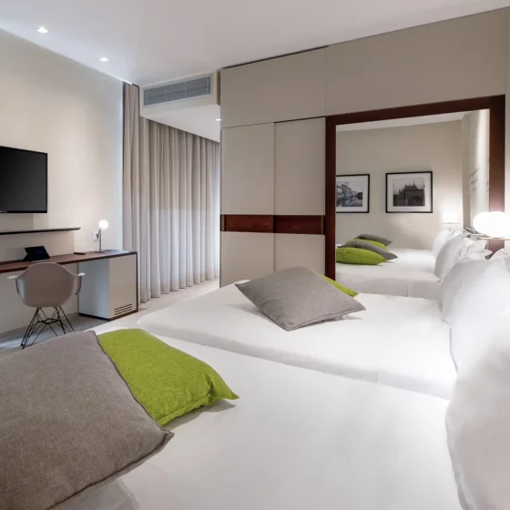 Hotel room with two twin beds in the foreground, gray pillows and green cushions, and a desk with a wall-mounted flat-screen TV.