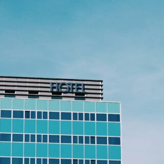 Partial view of a modern glass-front hotel against a blue sky, with the word HOTEL visible on the top edge.
