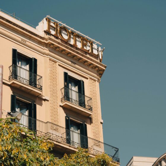 Beige European hotel/apartment building with decorative stonework and wrought-iron balconies, rooftop sign reading 'HOTEL' against a clear blue sky, with green shutters on the windows.