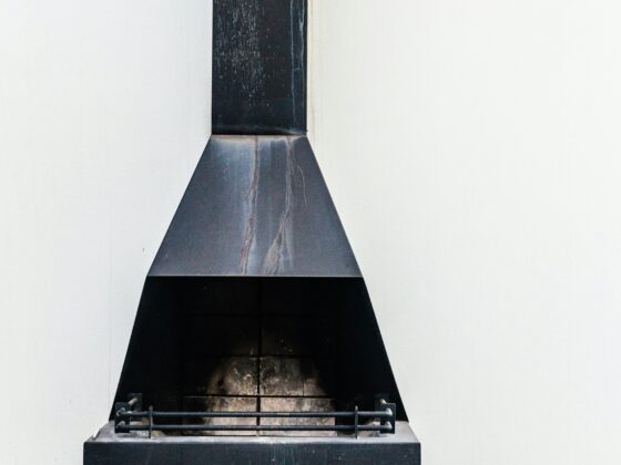 Outdoor black metal fireplace with a tall, narrow chimney and a wide, pyramid-shaped hood; logs sit nearby on a white patio wall backdrop.
