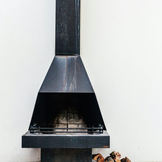 Outdoor black metal fireplace with a tall, narrow chimney and a wide, pyramid-shaped hood; logs sit nearby on a white patio wall backdrop.
