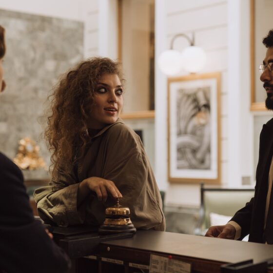 Woman with curly hair converses with two men in suits at a hotel reception desk, a bell visible on the counter.