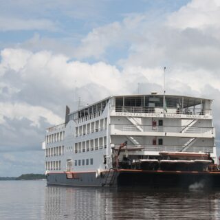 White cruise ship on a calm river under a partly cloudy sky; reads 'IBEROSTAR GRAND AMAZON' on the side, Manaus, Brazil.