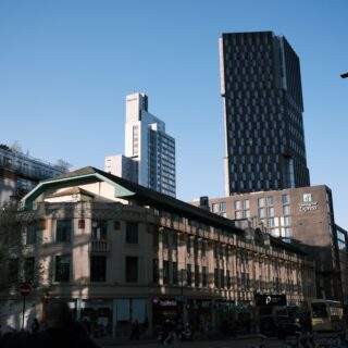 City street scene with a tall dark glass skyscraper and lighter mid-rise buildings against a clear blue sky; Holiday Inn Express sign visible on the foreground building.