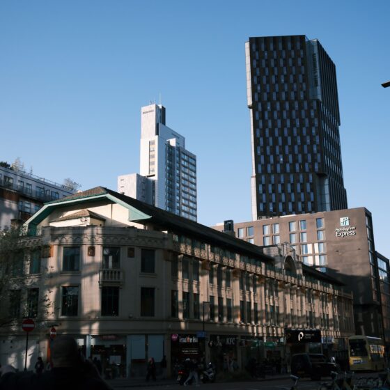 City street scene with a tall dark glass skyscraper and lighter mid-rise buildings against a clear blue sky; Holiday Inn Express sign visible on the foreground building.
