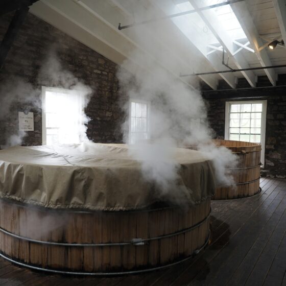 Inside a stone-walled brewery with a large steaming wooden fermentation vat under skylights.