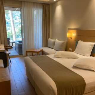 Hotel room with two large beds, neutral decor, bedside lamp, and a balcony door with sheer and heavy curtains revealing trees outside.