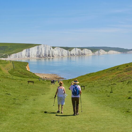 Hikers walking along a grassy coastal path toward white chalk cliffs and a calm blue sea under a clear sky.