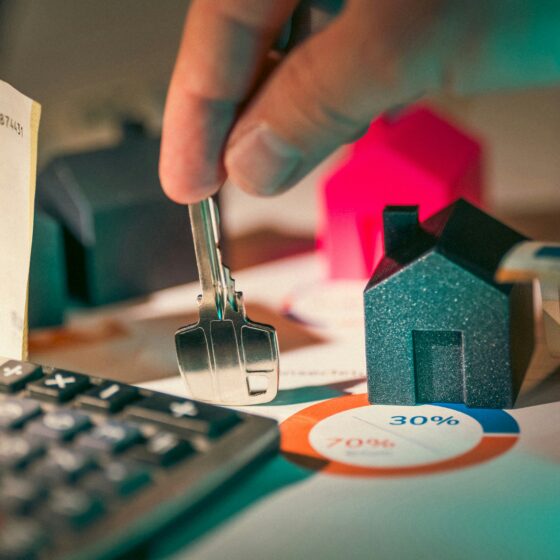Close-up of a hand holding a key near small house models on a desk, with a calculator and a percentage chart in view, suggesting financial planning or a mortgage.