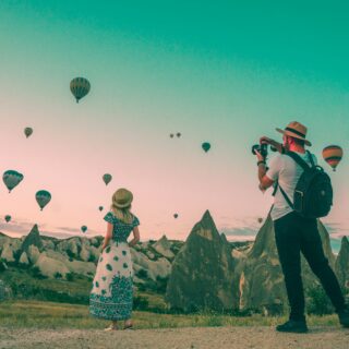 Two visitors on a rocky terrain watching dozens of colorful hot air balloons fill the sky; a man with a camera and backpack photographs while a girl in a patterned dress stands nearby.
