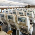 Inside a wide airplane cabin with rows of beige seats and personal entertainment screens on the back of each headrest, lights overhead.