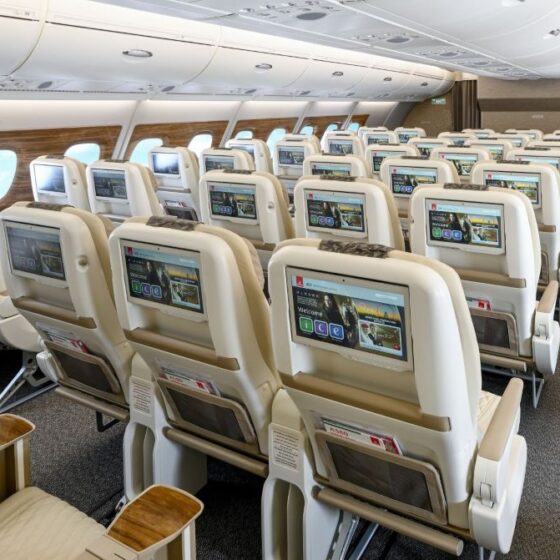 Inside a wide airplane cabin with rows of beige seats and personal entertainment screens on the back of each headrest, lights overhead.
