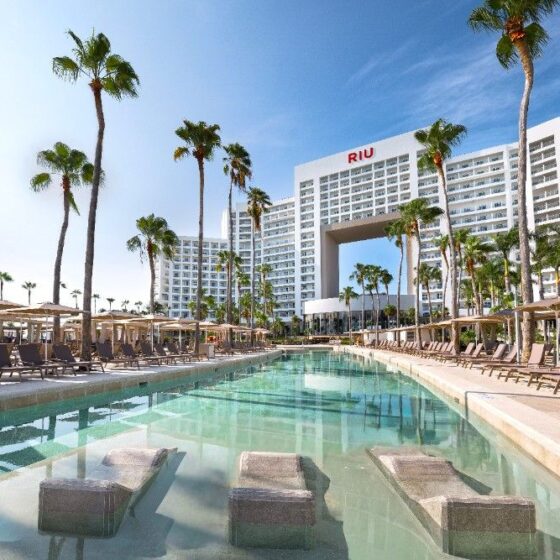 RIU hotel with a long pool surrounded by palm trees, umbrellas, and lounge chairs on a sunny day while guests relax nearby.