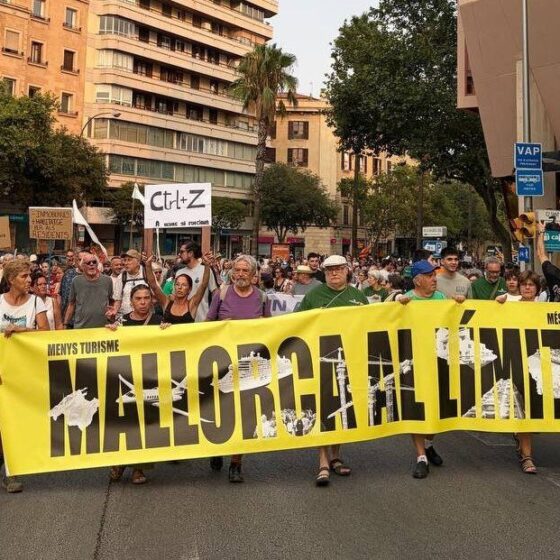 Crowd of protesters marching down a city street, holding a bright yellow banner that reads 'MALLORCA AL LÍMIT!' with smaller signs in the background and onlookers watching nearby.