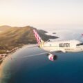 Volotea passenger jet flying over a coastal landscape with beach, hills, and sea at sunset/soft light.