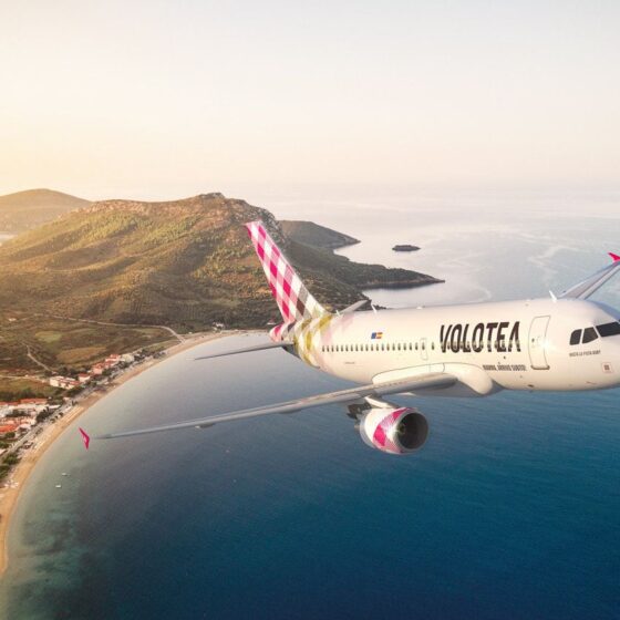 Volotea passenger jet flying over a coastal landscape with beach, hills, and sea at sunset/soft light.
