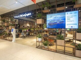 Interior of a travel agency with a 'Viajes El Corte Ingles' sign, desks, and seating under warm lighting, plus a large cruise advertisement on a screen.