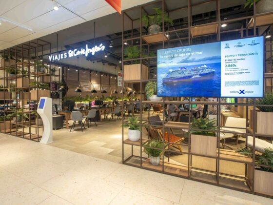 Interior of a travel agency with a 'Viajes El Corte Ingles' sign, desks, and seating under warm lighting, plus a large cruise advertisement on a screen.