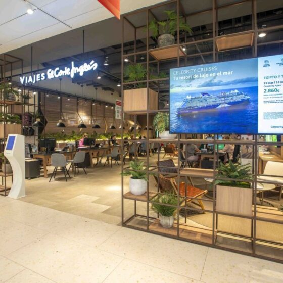 Interior of a travel agency with a 'Viajes El Corte Ingles' sign, desks, and seating under warm lighting, plus a large cruise advertisement on a screen.