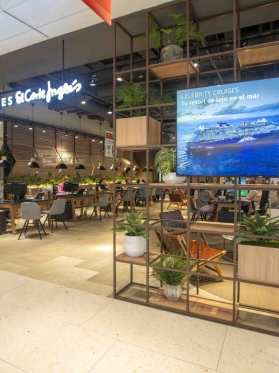 Interior of a travel agency with a 'Viajes El Corte Ingles' sign, desks, and seating under warm lighting, plus a large cruise advertisement on a screen.