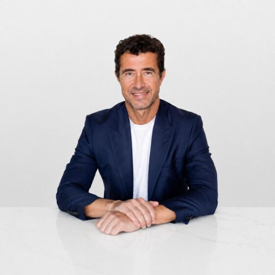 Portrait of a man in a navy blazer seated at a white table, smiling at the camera against a light background.