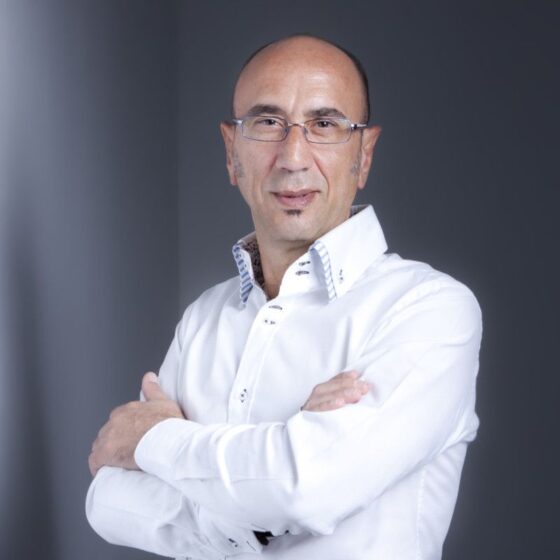 Professional portrait of a man with glasses wearing a white dress shirt, arms crossed, against a gray gradient background.