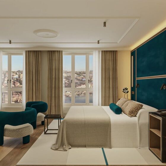 Luxurious hotel room with a teal accent wall, large bed, beige bedding, and turquoise chairs facing a window view of the cityscape.