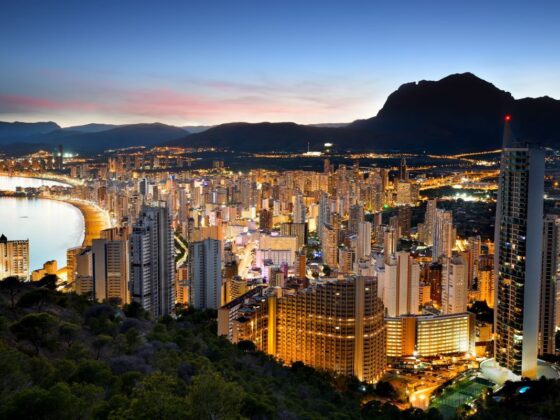 Coastal city skyline at dusk with lit high-rise buildings along a curved beach and mountains in the background. Note: This conveys the scene's main elements and mood without overloading details.