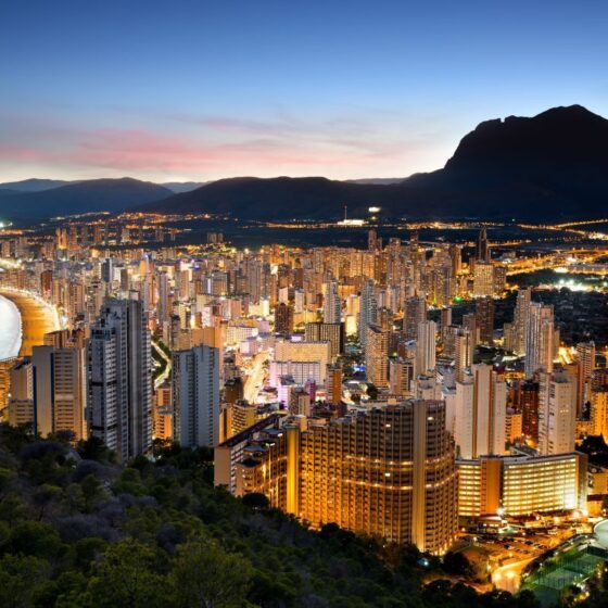 Coastal city skyline at dusk with lit high-rise buildings along a curved beach and mountains in the background. Note: This conveys the scene's main elements and mood without overloading details.