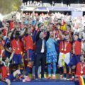 Spain national football team celebrates victory with trophy on stage, players wearing red and blue jerseys and gold medals