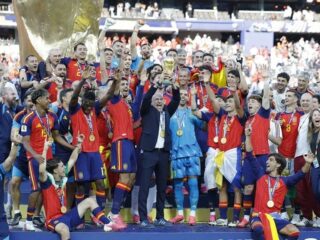 Spain national football team celebrates victory with trophy on stage, players wearing red and blue jerseys and gold medals