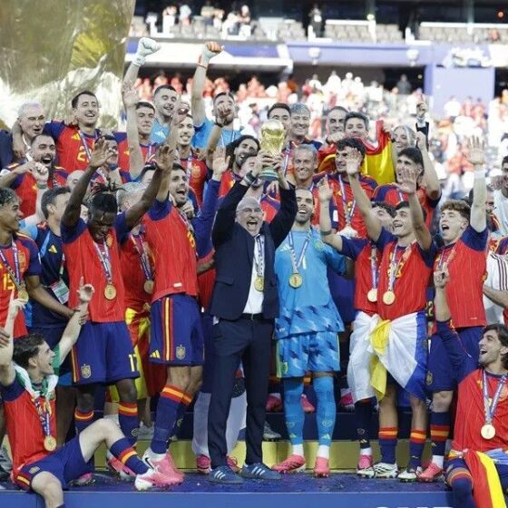 Spain national football team celebrates victory with trophy on stage, players wearing red and blue jerseys and gold medals