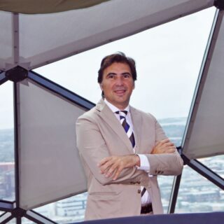 Confident businessman in a beige suit with arms crossed, standing inside a glass-framed atrium overlooking a city.
