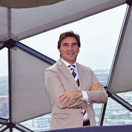 Confident businessman in a beige suit with arms crossed, standing inside a glass-framed atrium overlooking a city.