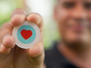 Close-up of a person pinching a round button with a red heart inside a blue circle, like/thumbs-up symbol in focus, blurred background.