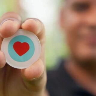 Close-up of a person pinching a round button with a red heart inside a blue circle, like/thumbs-up symbol in focus, blurred background.