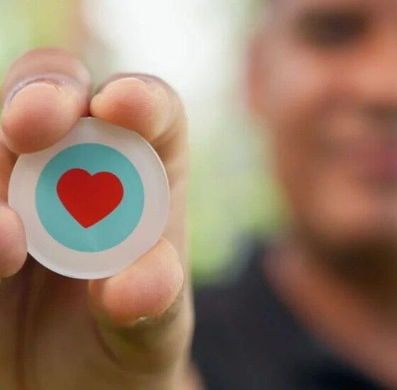 Close-up of a person pinching a round button with a red heart inside a blue circle, like/thumbs-up symbol in focus, blurred background.