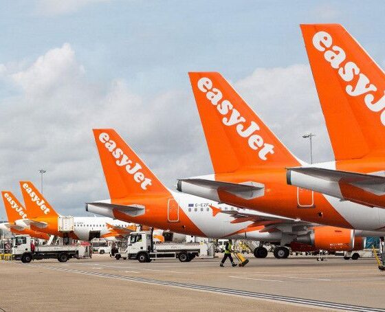Multiple bright orange easyJet airplanes are parked at a busy tarmac with ground support vehicles nearby.