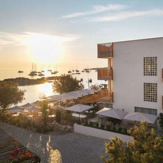 Sunset over a resort by the sea with white building, balconies, and outdoor seating under umbrellas facing calm water and boats